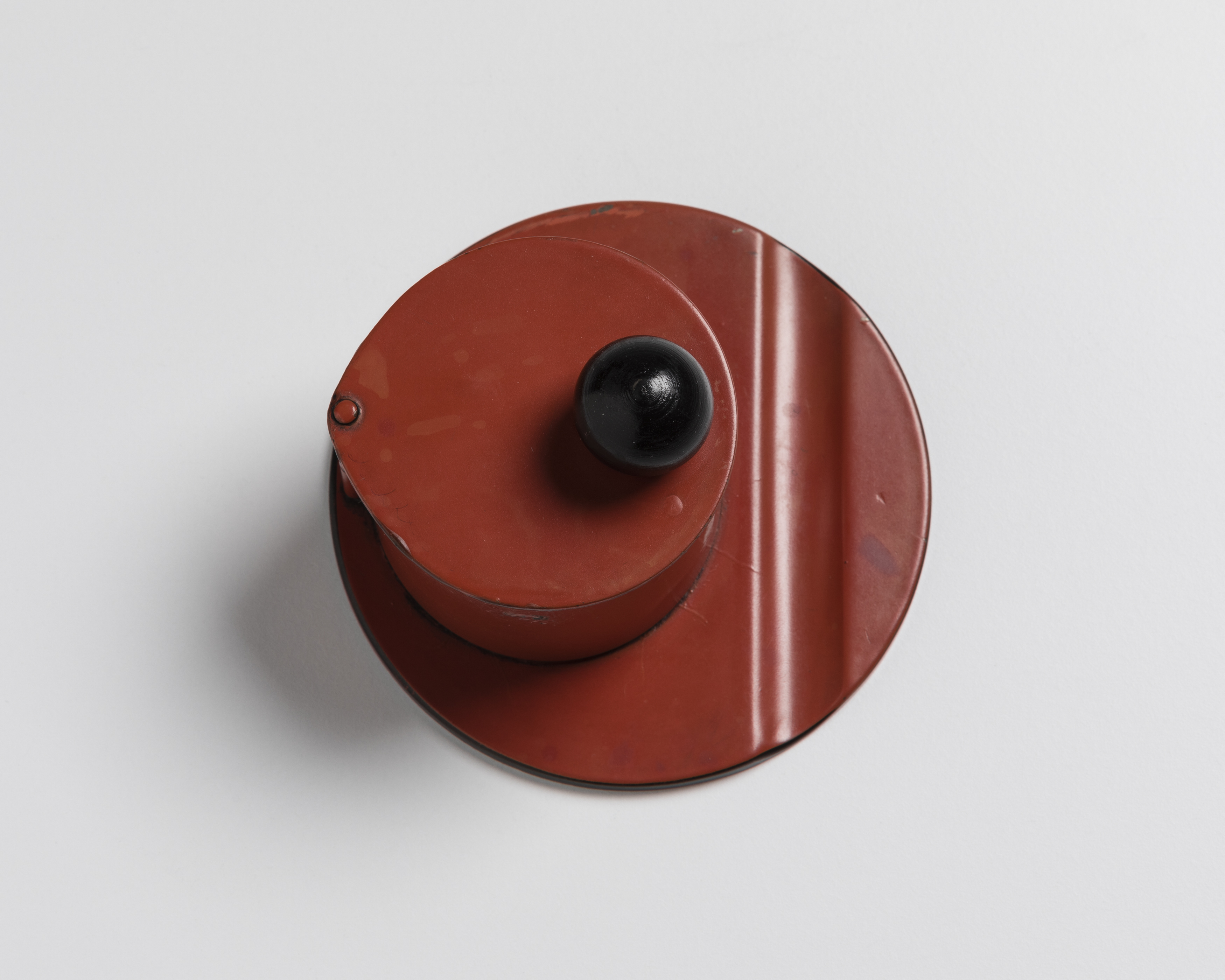 Red circular inkstand with a black ball finial on hinged cover, photographed from above.